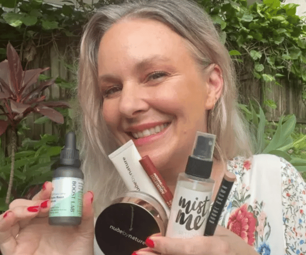 Blonde woman smiling, holding up several beauty products.