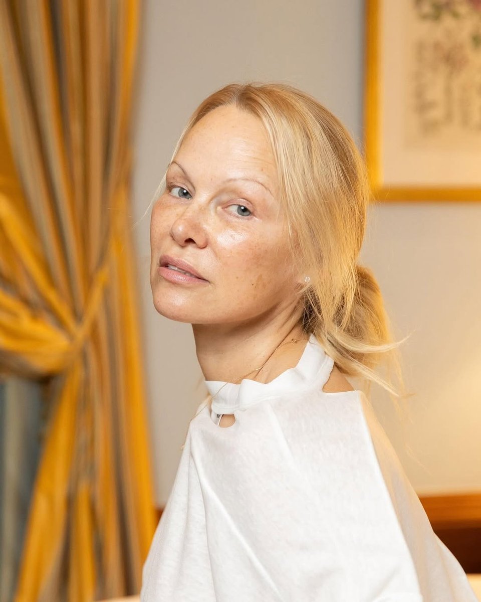 Pamela Anderson poses for the camera with her hair tied back in a low bun, wearing a white shirt.