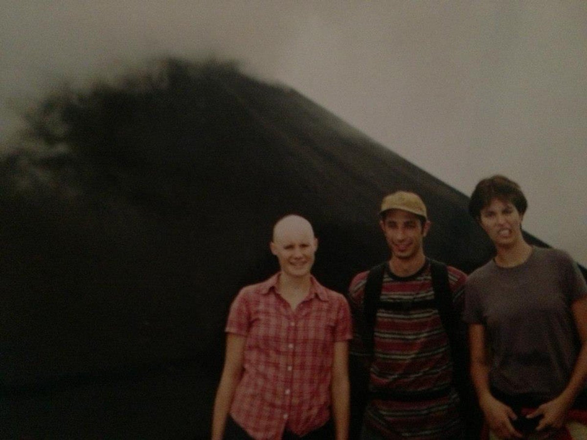 A bald woman wearing a check shirt poses confidently outdoors with two friends.