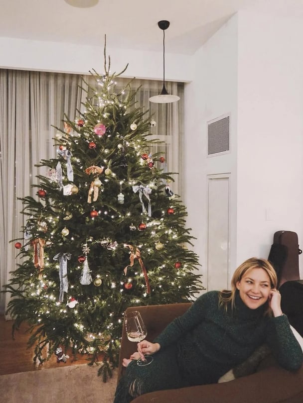 Kate Hudson in a green outfit, holding a glass of wine and sitting infront of her Christmas tree.