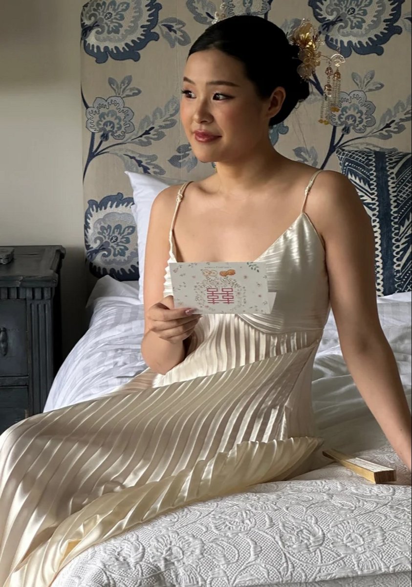 Woman wearing bridal gown, sitting on a bed, holding a card.