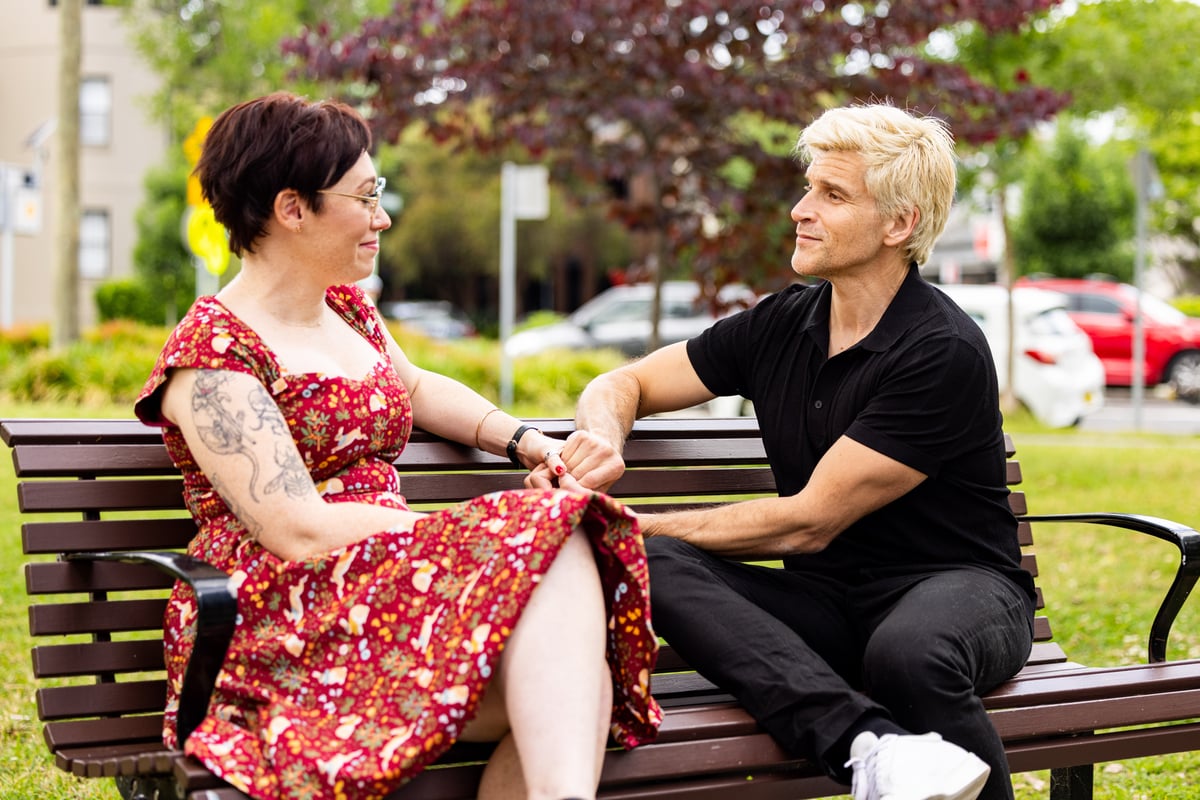 A woman with short brown hair wearing a red dress sits on a park bench next to Osher Gunsberg dressed in black.