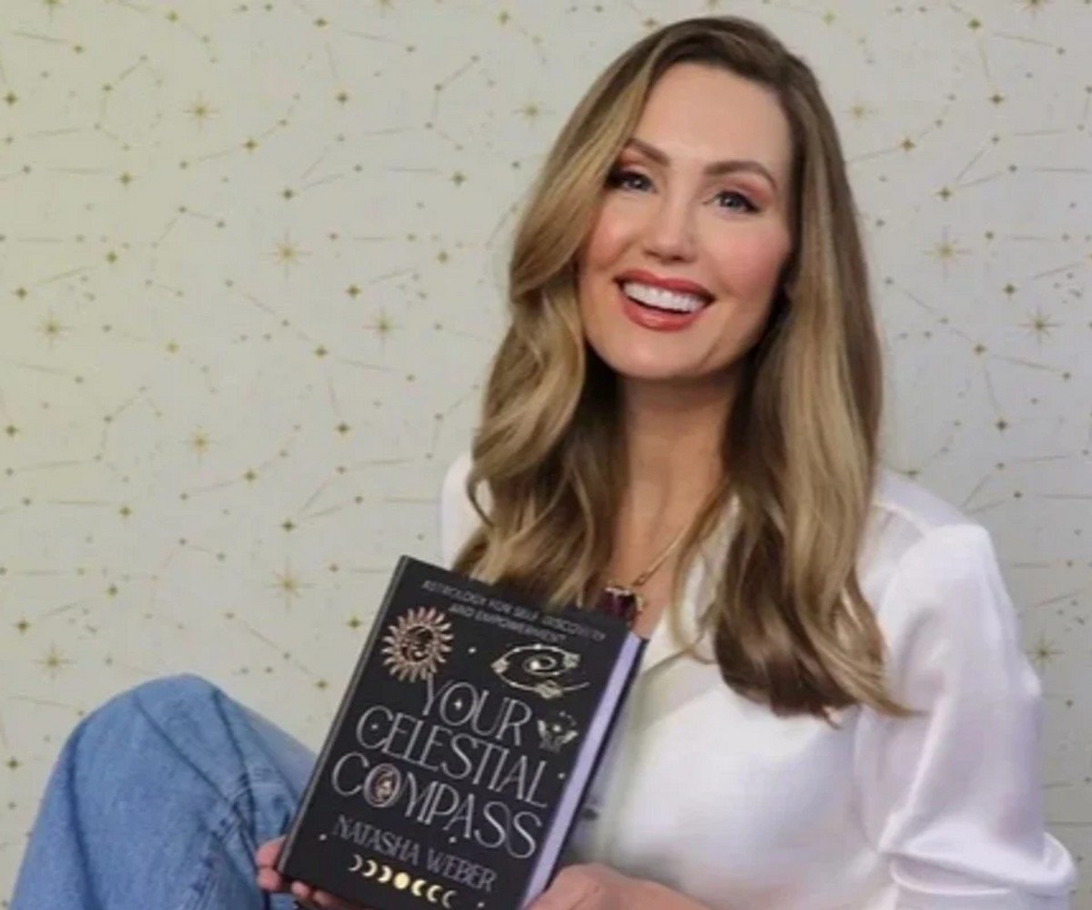 Natasha Weber holding her book Your Celestial Compass. Blonde woman wearing jeans and white blouse, sitting down holding a book.