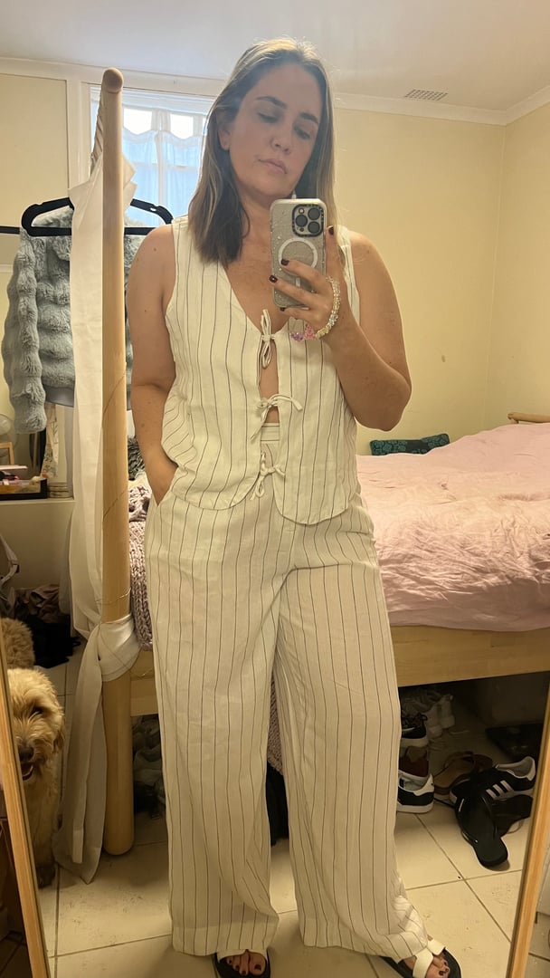 Woman wears white linen matching top and pants with pinstripes, standing in a bedroom.