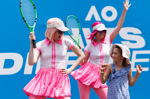 Kids playing on the AO tennis court with some Tennis Australia performers wearing pink wigs.