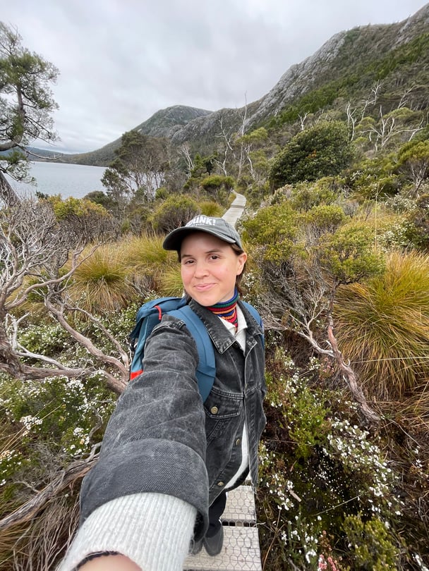 A selfie of a woman with brown hair in bushland, hiking in a black denim jacket and blue backpack.