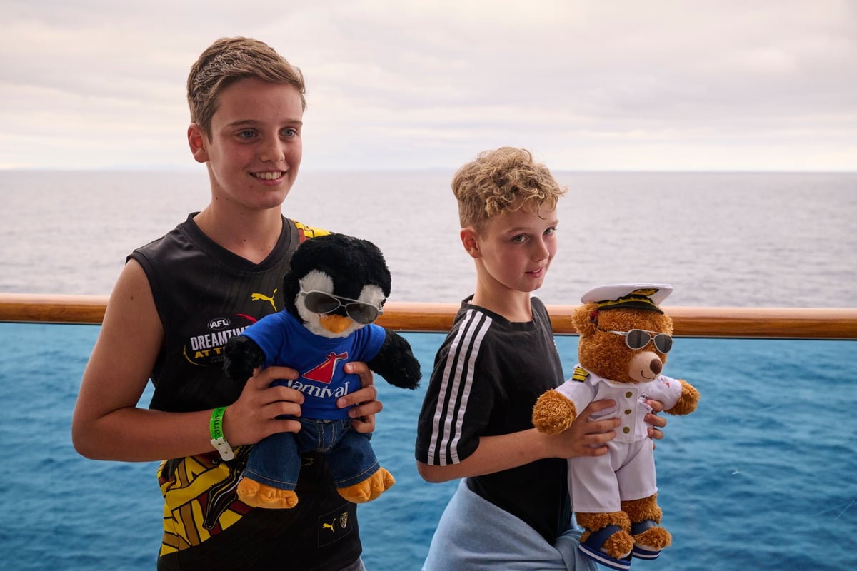 Two young boys holding a toy bear and a toy penguin with the sea in the background.