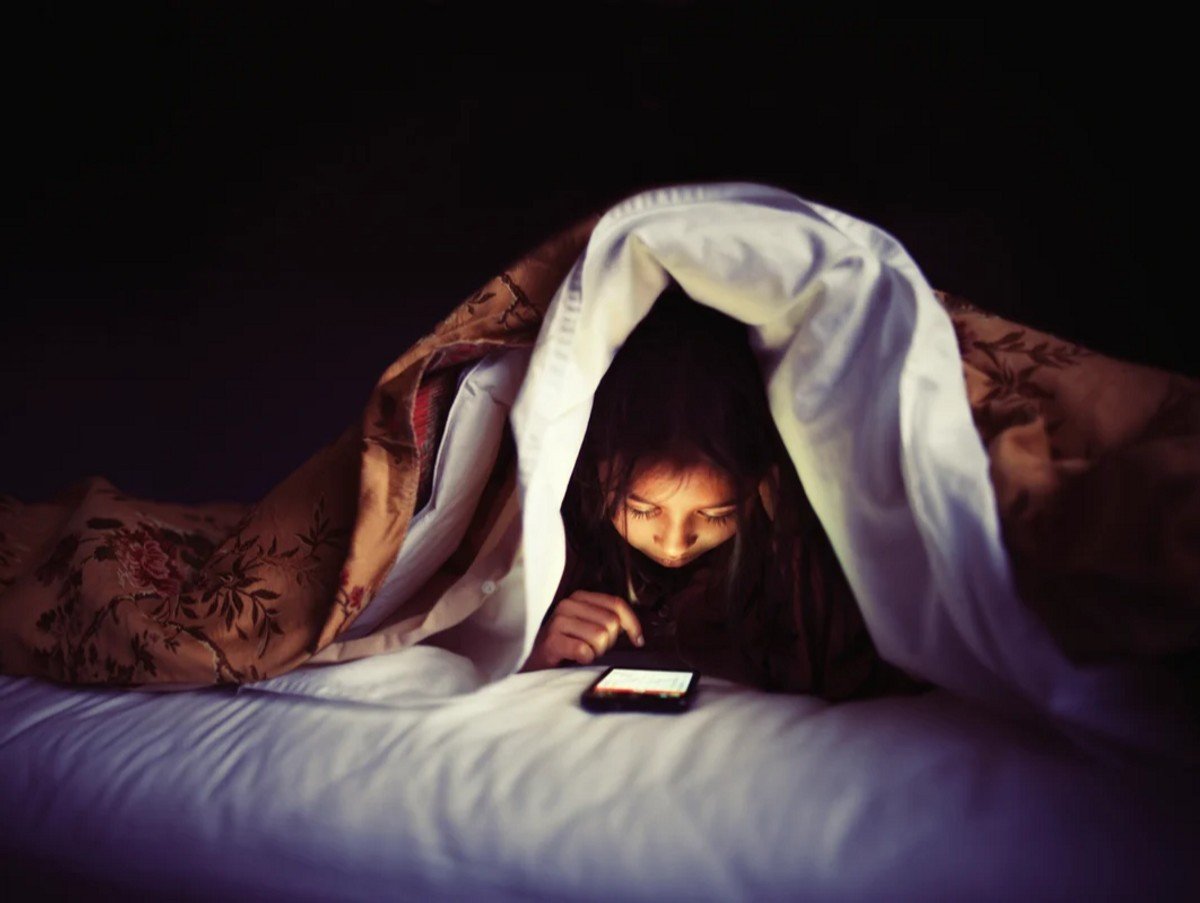 Girl using smartphone under bed covers.