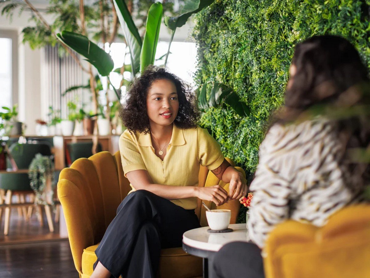 Two people engage in a pleasant conversation, sitting in comfortable yellow armchairs in a cafe with lush greenery.