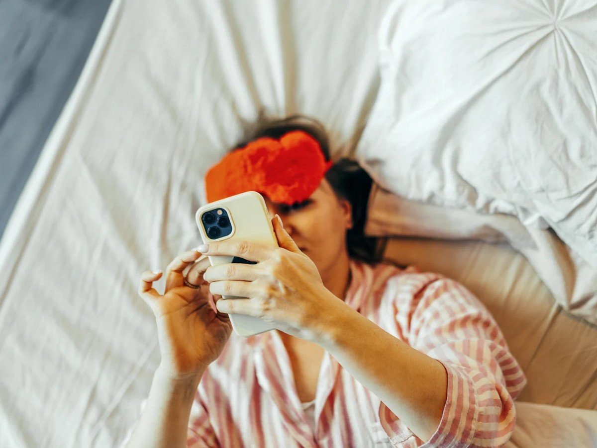 A woman in the bed with red Eye Mask and mobile phone in hand.