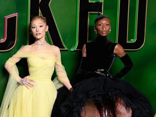 Ariana Grande in a yellow gown and Cynthia Erivo in a black gown, posing in from of a Wicked sign on the red carpet.