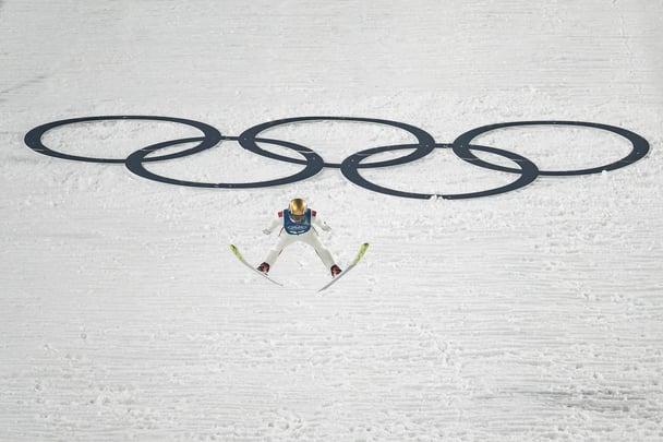 A ski jumper with his legs spread as he jumps over snow, with large Olympic rings behind him.