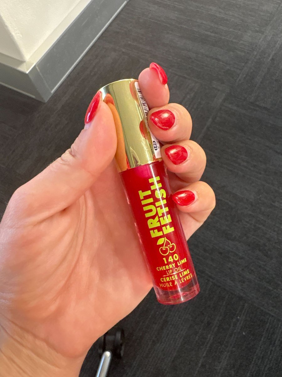 A picture of Milani Fruit Fetish Lip Gloss held in a hand with a red manicure.