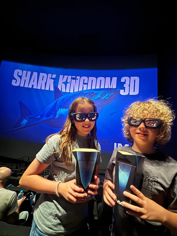 Two tweens wearing 3D glasses holding popcorn in front of the seven-storey screen inside the IMAX Melbourne.
