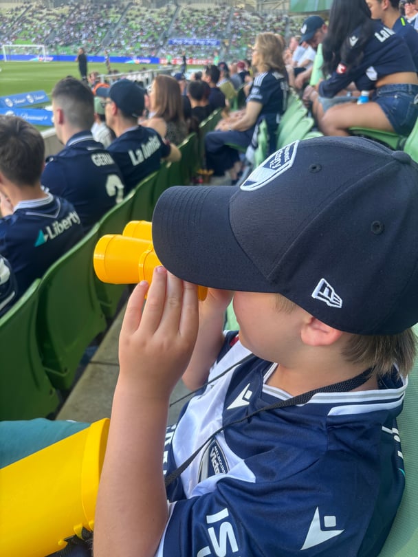 The writer's son seated in the stadium overlooking the grounds through yellow kids' binoculars.