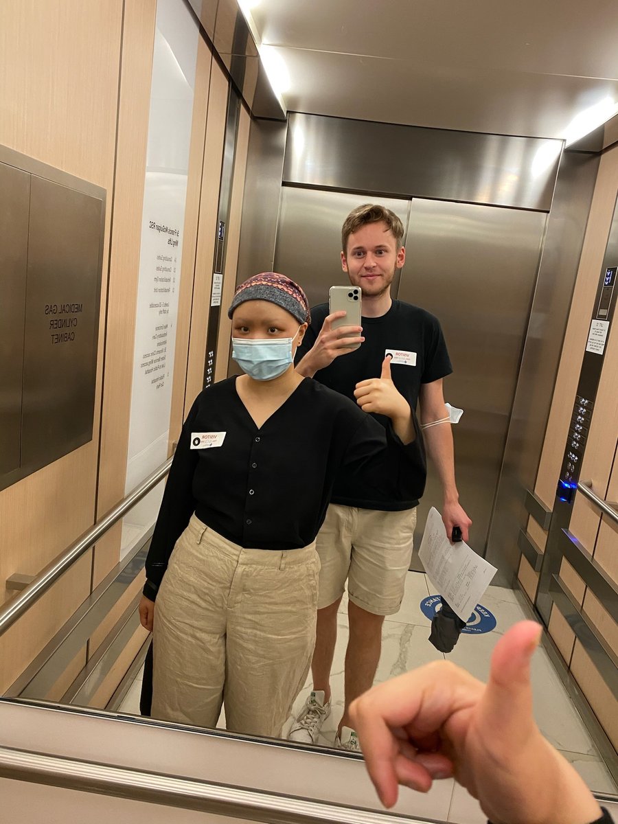 A woman wearing a face mask and beanie stands in front of a man taking a mirror selfie in an elevator.