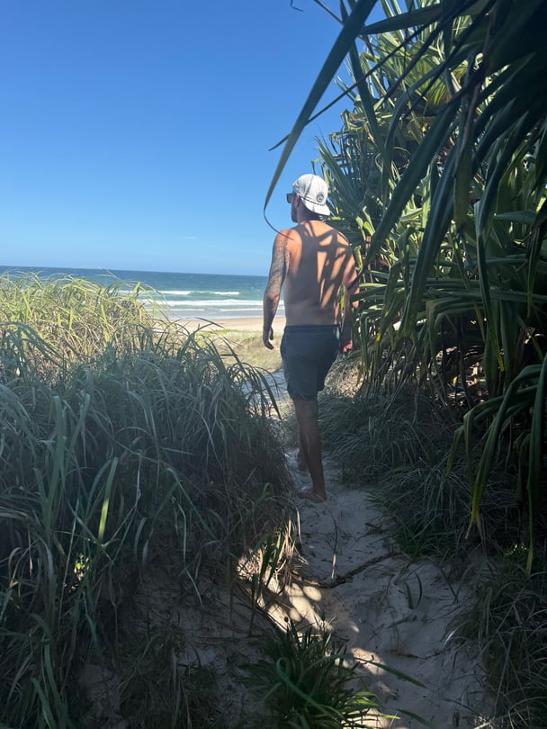 A man walks through a small entrance amongst bushes to a beach.