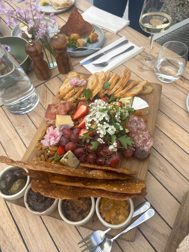 A charceturie board with cracked, breads, meats, grapes, strawberries, cheese and pink flowers.