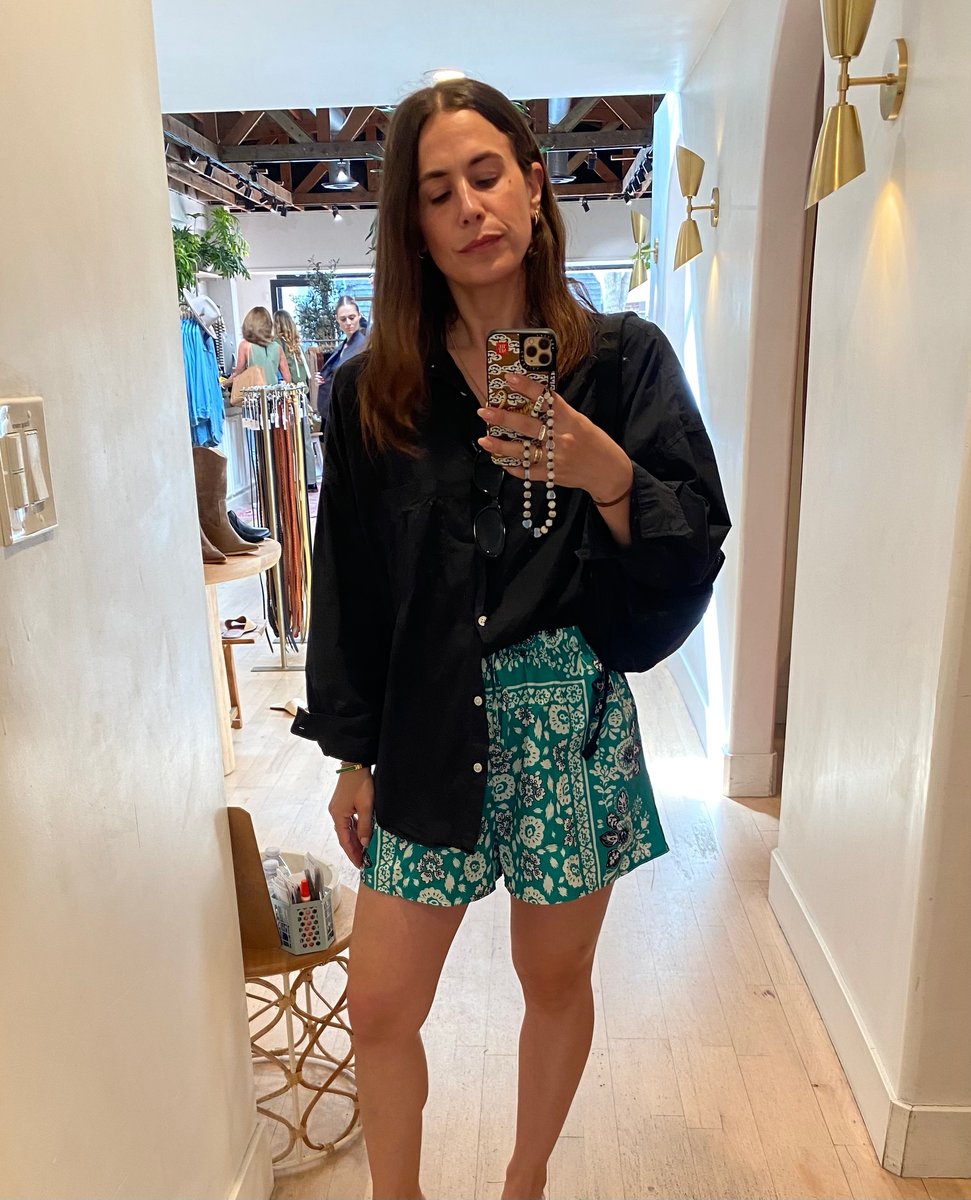 A woman takes a changeroom selfie in an oversized black shirt and printed shorts.