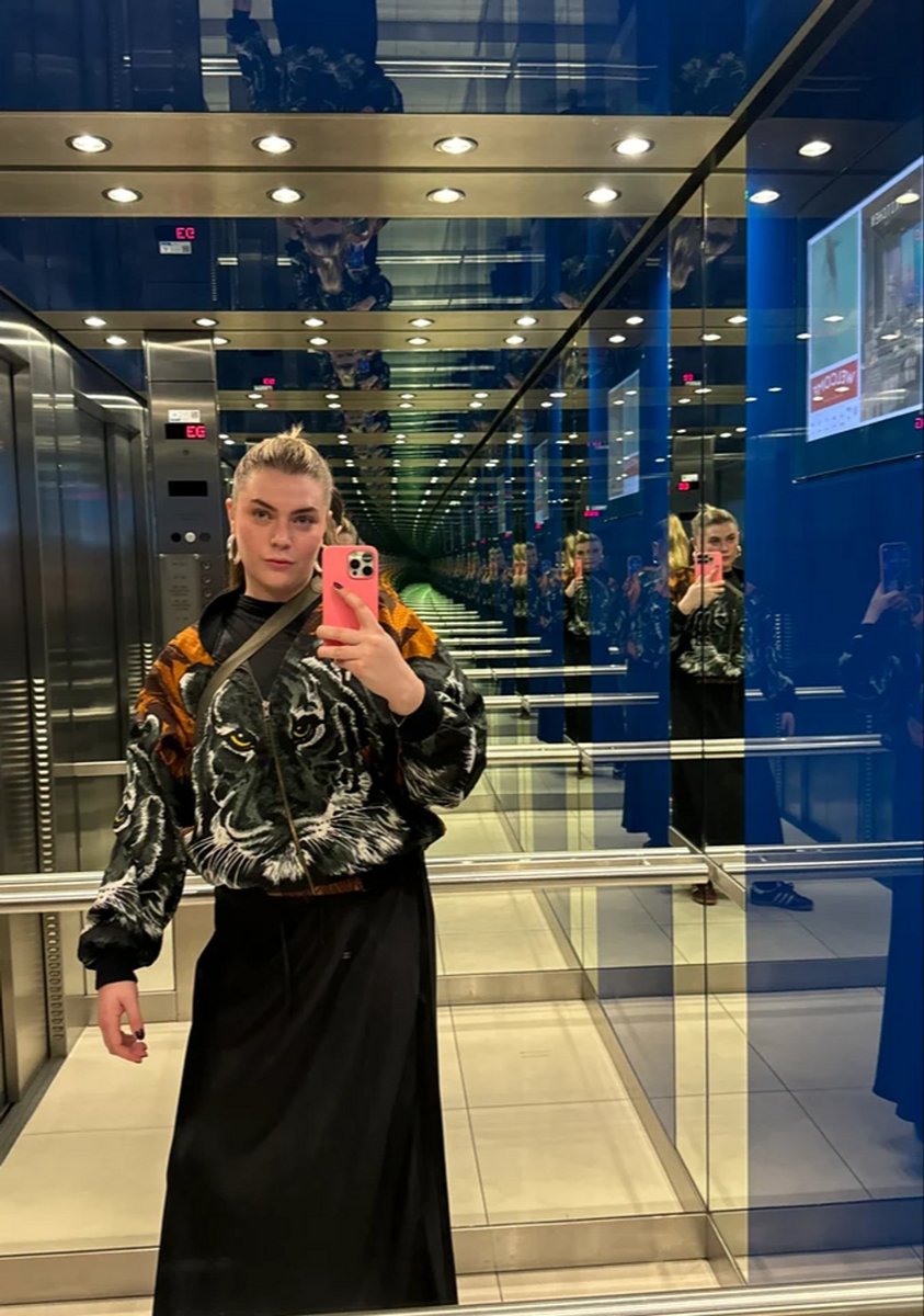 The journalist taking a mirror selfie in an elevator, wearing a long black skirt and tiger jacket. 