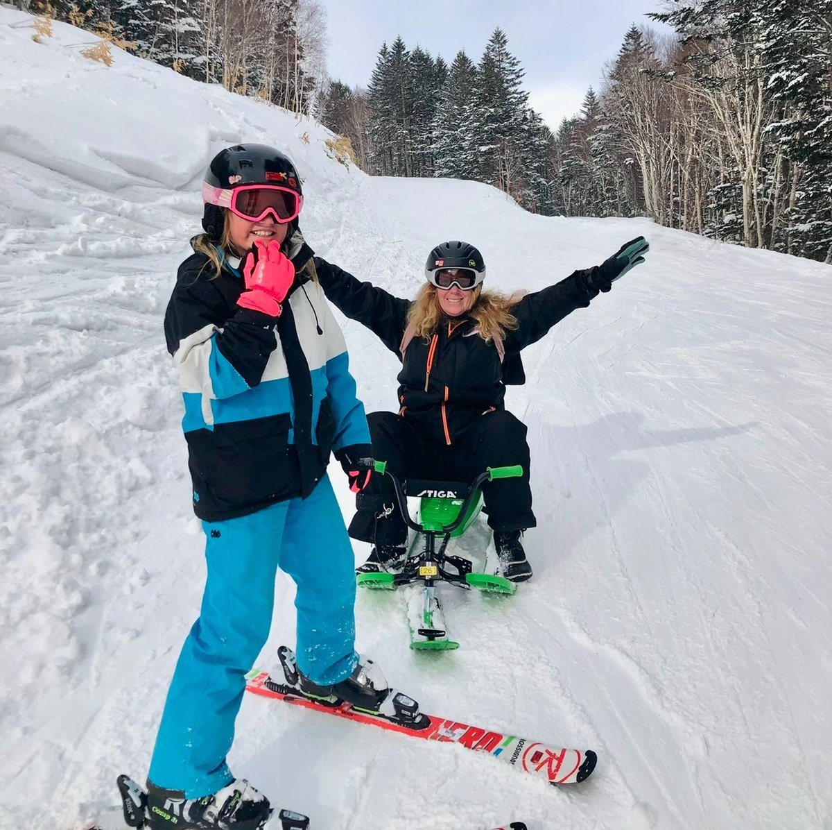 A mother and her young daughter ski side by side on a snowy slope. The mother raises her arm, cheering her child on.
