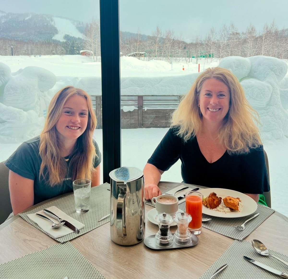 A mother and daughter sit at an outdoor café in a snowy setting, smiling at each other while eating lunch.