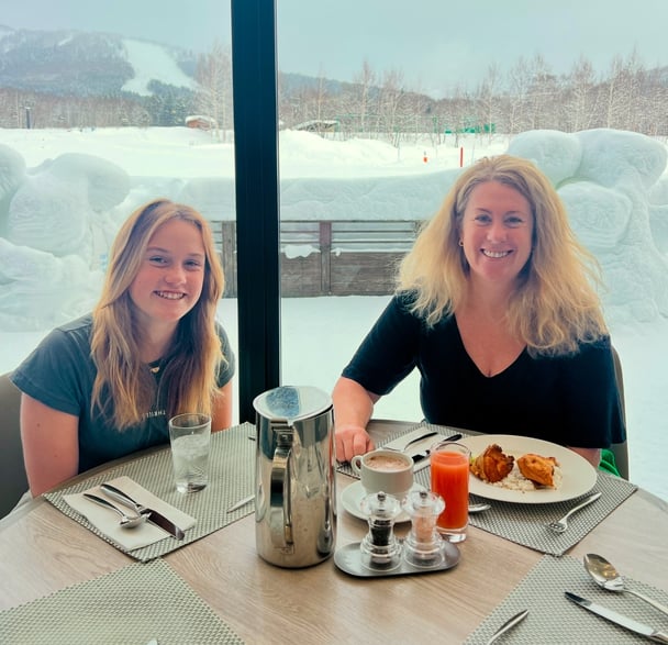 A mother and daughter sit at an outdoor café in a snowy setting, smiling at each other while eating lunch.