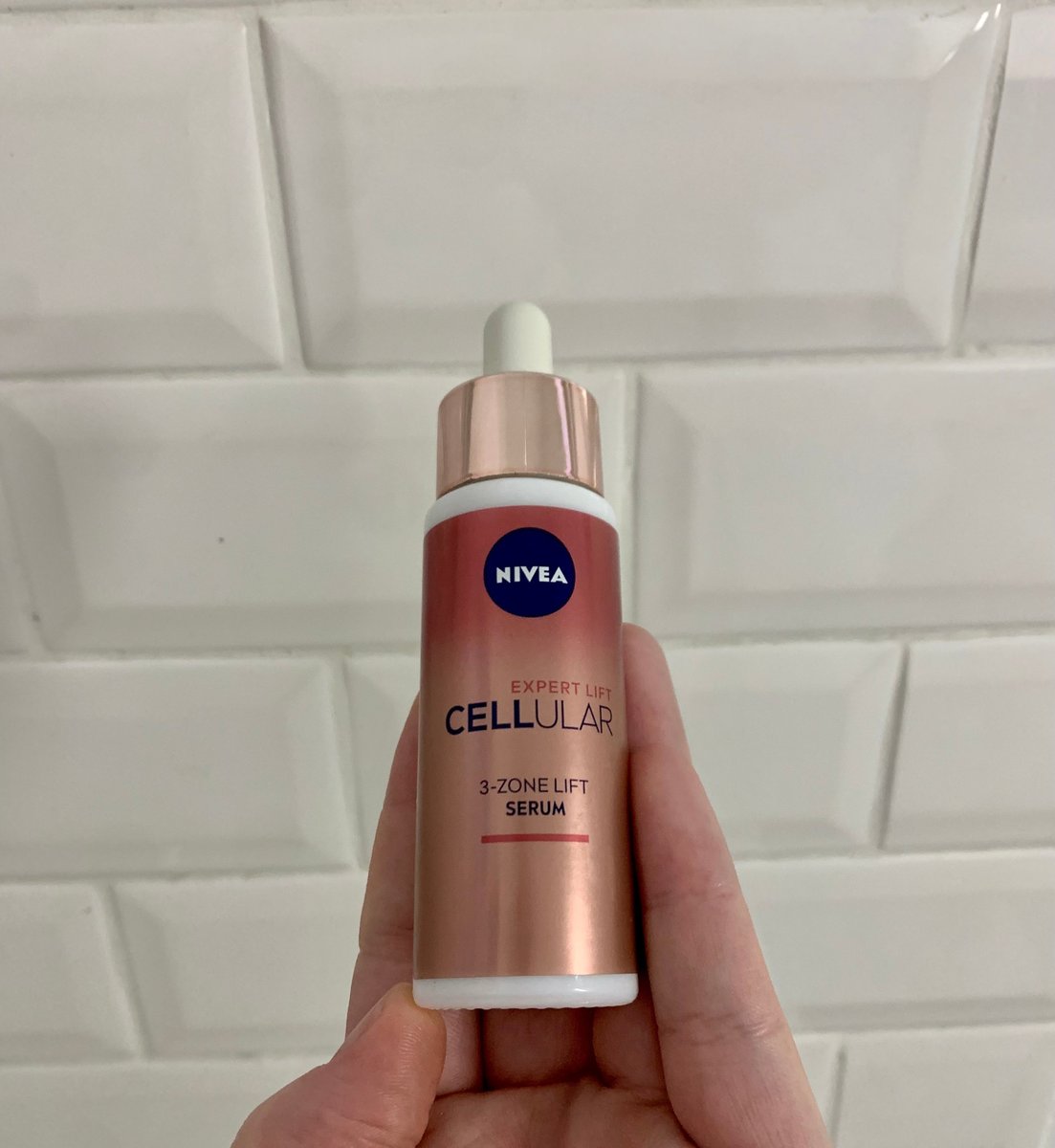 A hand holding Nivea Cellular 3 Zone Lift Serum against white bathroom tiles.