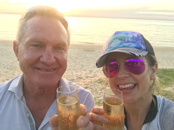 Father and adult daugher on a beach holding glasses of champagne. The woman is wearing a cap and reflective sunglasses.