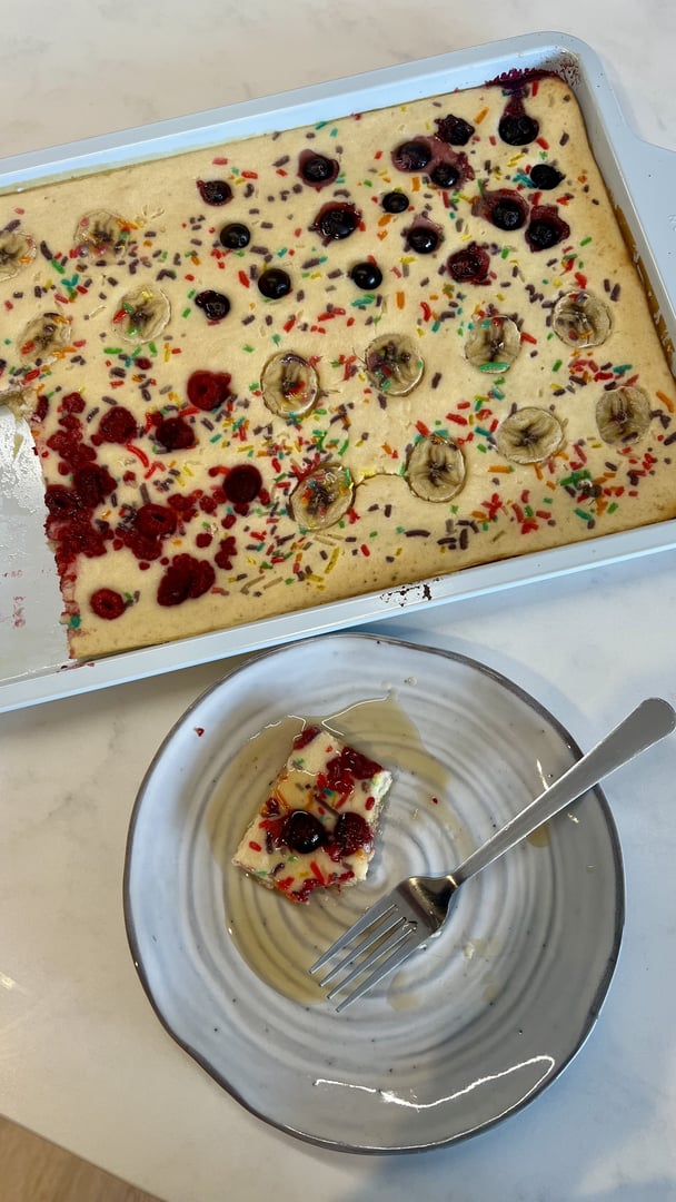 A rectangular tray of baked pancakes topped with sliced banana, mixed berries and rainbow sprinkles, with a single square slice served on a plate drizzled with honey and a fork beside it.