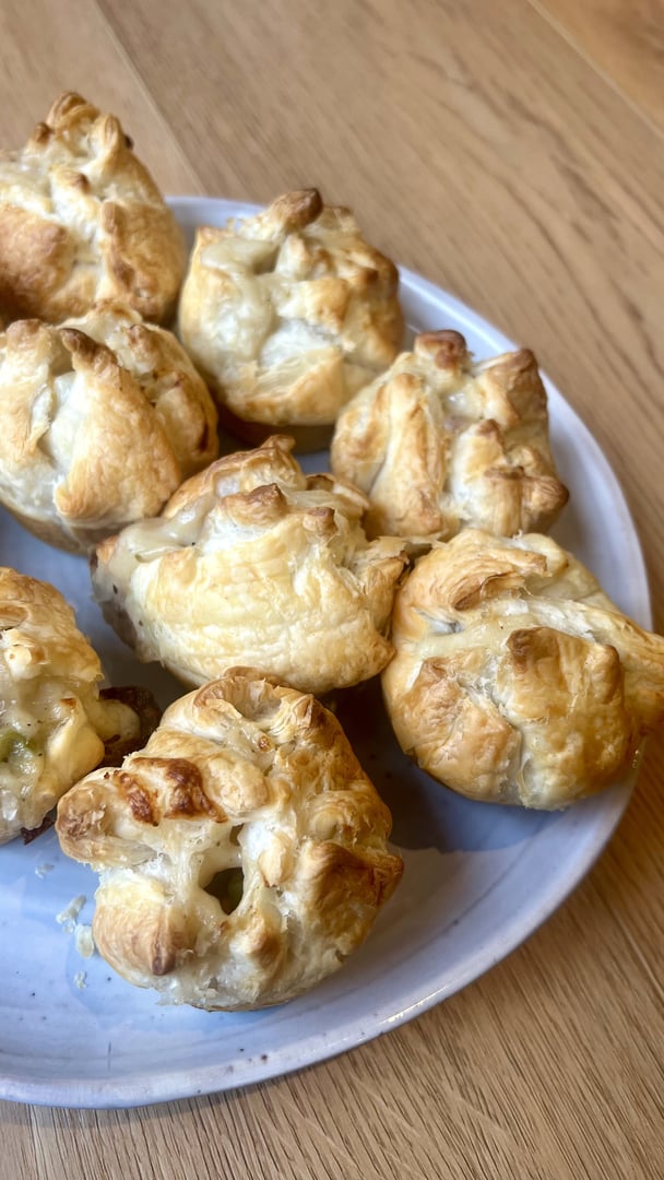 A plate of golden, flaky mini chicken pies made with puff pastry, baked until crisp and lightly browned, sitting on a white plate on a wooden table.