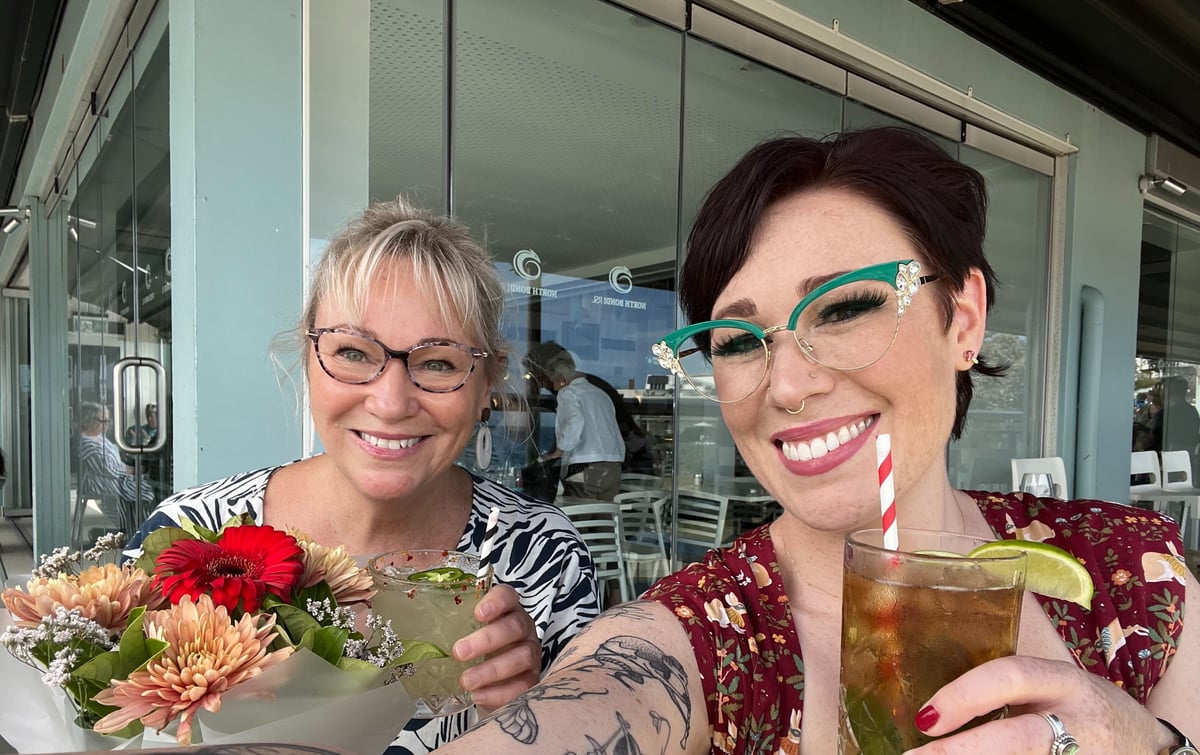 A blonde woman with glasses smiles holding flowers and a cocktail next to a smiling woman with short brown hair, who is also wearing glasses and holding drink.