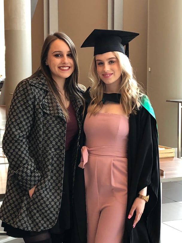 Krystal and Stephanie smiling together at graduation. Stephanie is in a pink dress with cap and gown. Krystal is in a patterned coat.