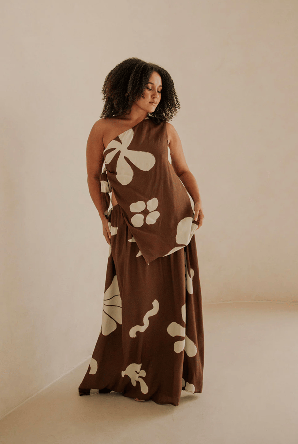 A brown maxi dress featuring one-shoulder detail, and an oversized white floral print.