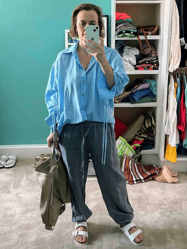 Mia Freedman wearing a blue shirt, grey linen trousers, and white Birkenstocks. She's also carrying a khaki-coloured leather tote.