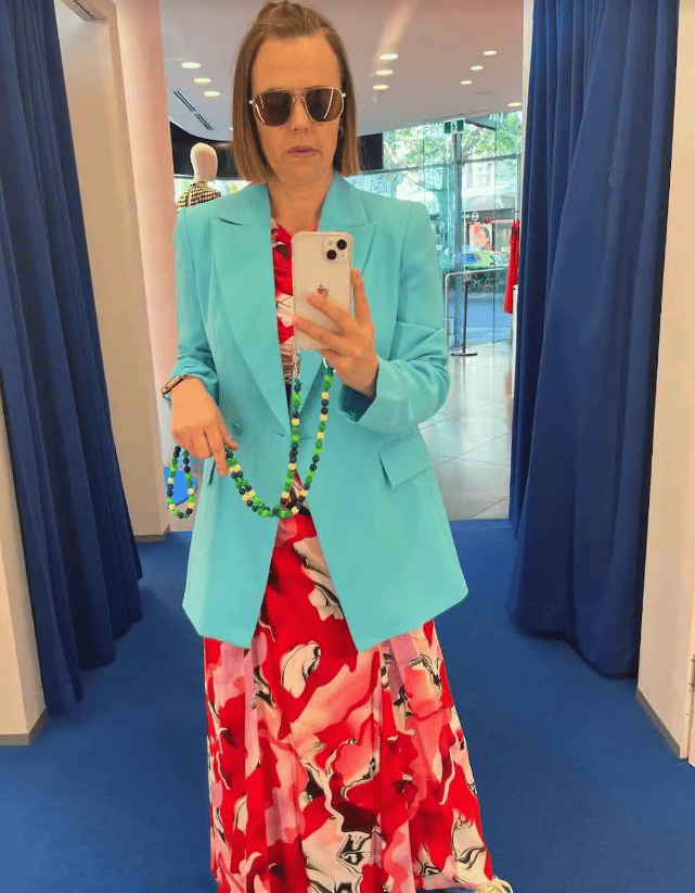 Mia Freedman taking a selfie in a change room wearing a red and pink floral floor-length dress, sneakers and a baby blue blazer. She's also wearing sunglasses and has a green beaded strap on her phone.