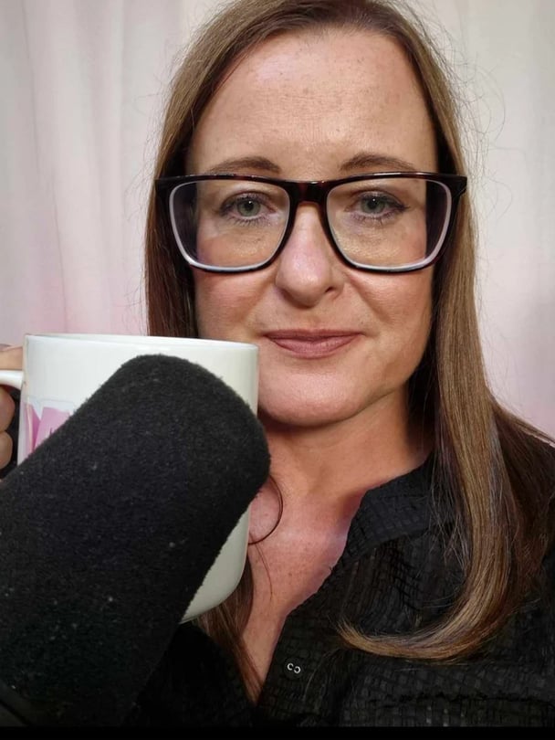 Woman with brown hair wearing black glasses and holding a coffee cup with a black microphone.