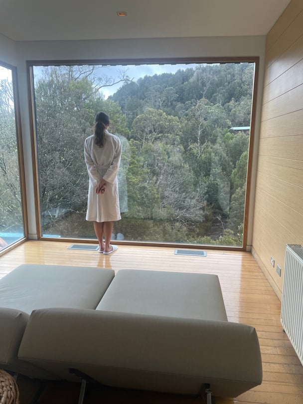 The back of a woman in a white robe and slippers, looking out the window onto bushland.