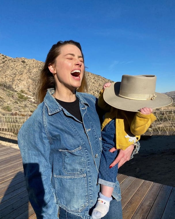 Amber Heard smiles with her young daughter Oonagh Paige, who has a wide-brimmed hat covering her face.