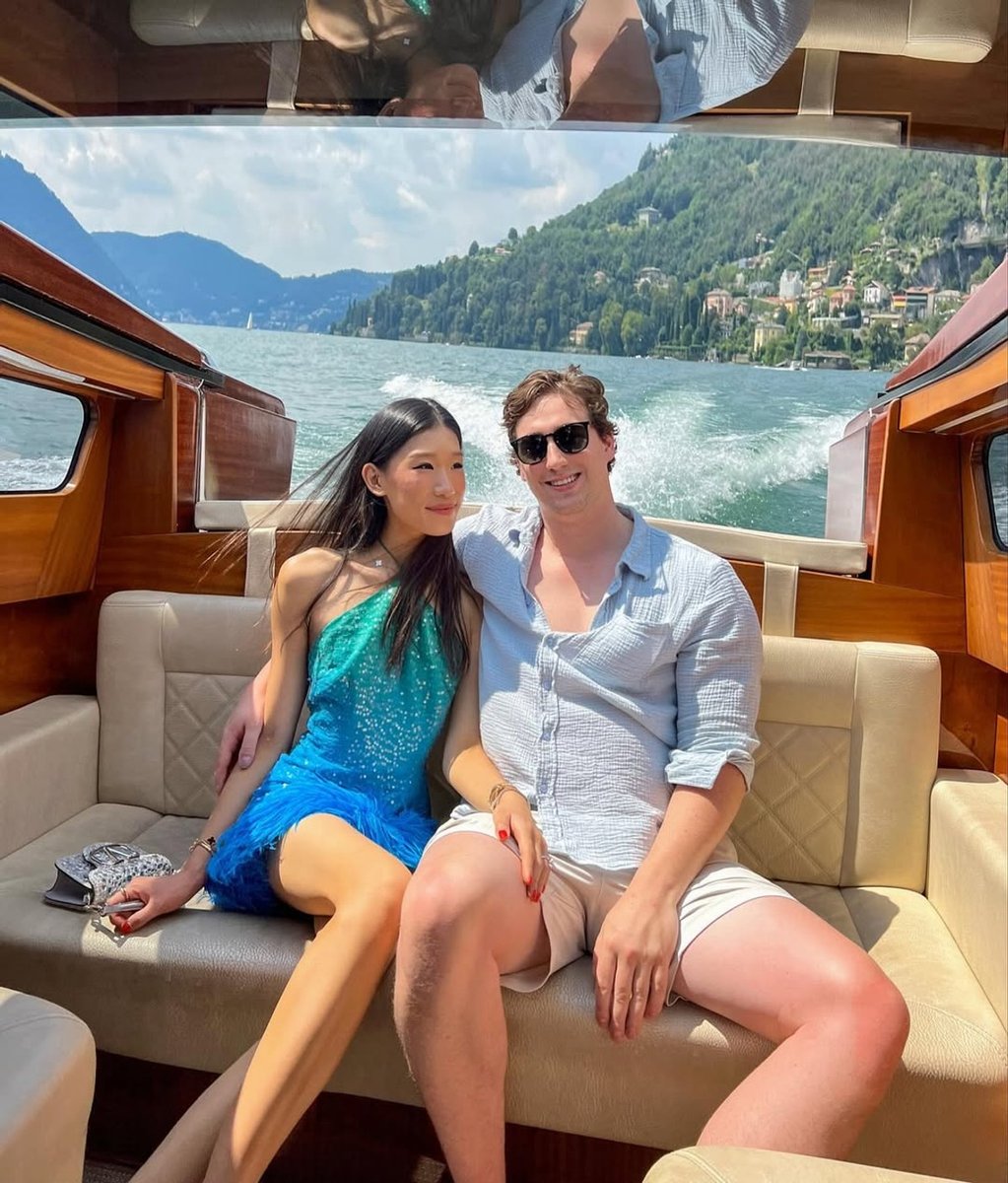 #RichTok influencer Rebecca Ma, aka Becca Bloom, and her husband David Pownall smile on a bought in Europe. Becca wears a sparkly blue halter dress with a feathered trim while David wears sunglasses, a light blue linen button down and cream shorts.