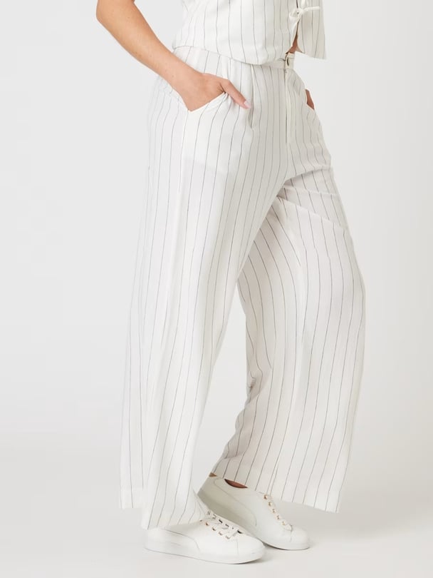 Bottom half of a woman's body, wearing white linen pants with a wide pinstripe, and white sneakers.