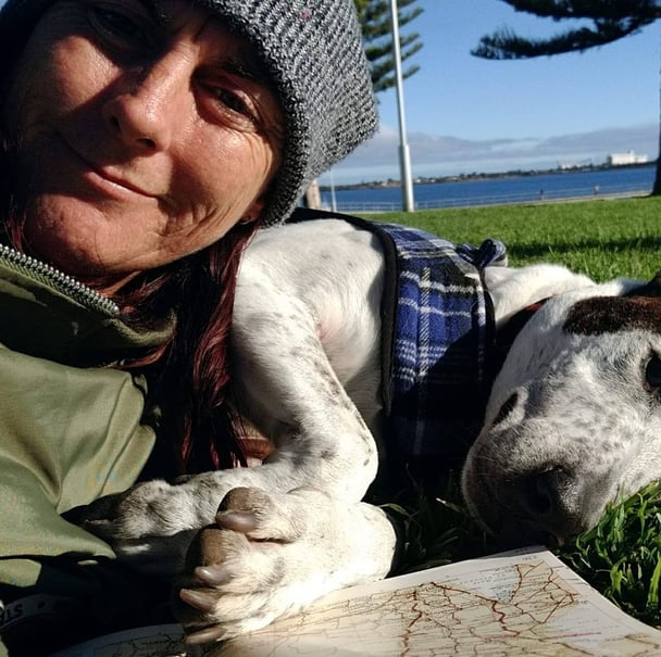 A smiling woman in a beanie lies on the grass next to her dog.