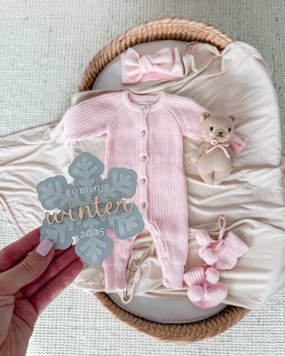 A flat lay of baby girl's pink clothing with a woman's hand holding a 'coming winter 2025' snowflake sign.