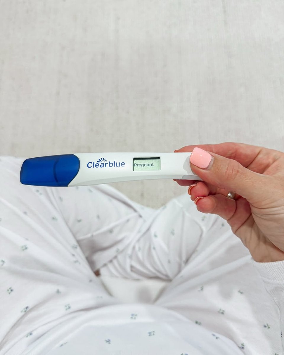 Caitlin Bailey holds a positive Clear Blue pregnancy test above her lap while in white pyjamas.