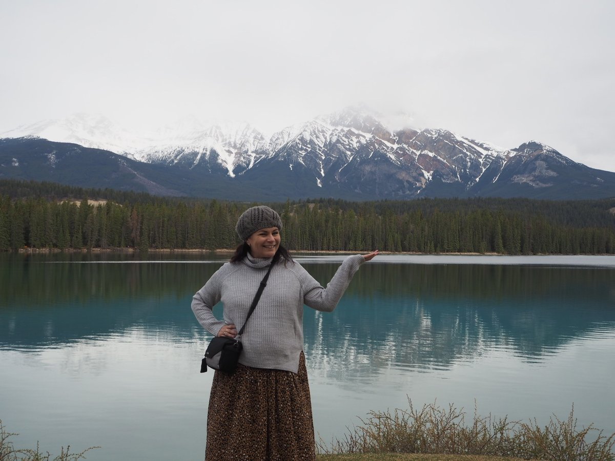 Woman wearing a grey turtleneck jumper and a textured maxi skirt poses in front of a lake and snow-capped mountains.