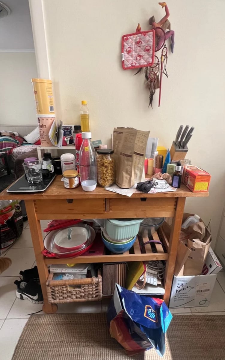 A very messy, disorganised kitchen. Clutter sits atop a bench on a kitchen island.