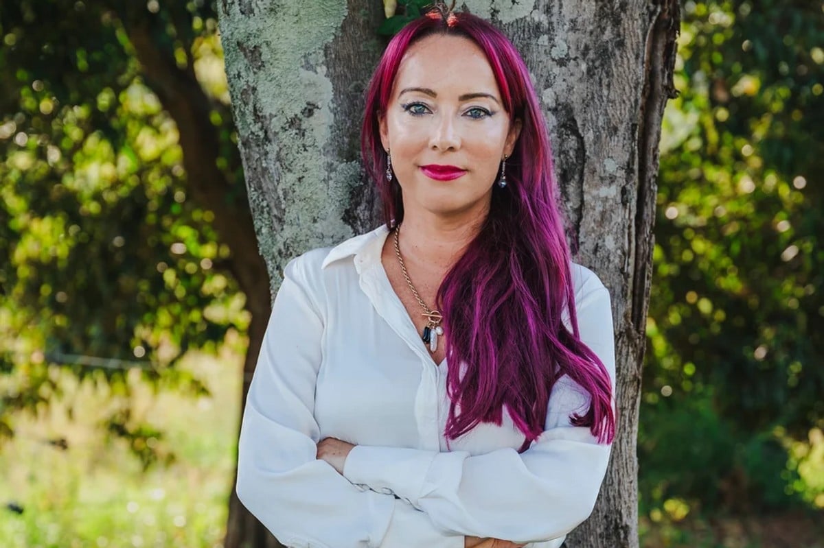 Anti-domestic violence advocate and mother Jas Rawlinson smiles with her arms crossed while standing in front of a tree.