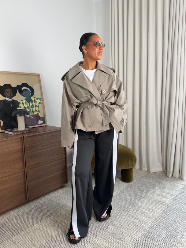 Emelia stands in black pants with a white stripe and a short tan trench coat, with sunglasses and a bun.