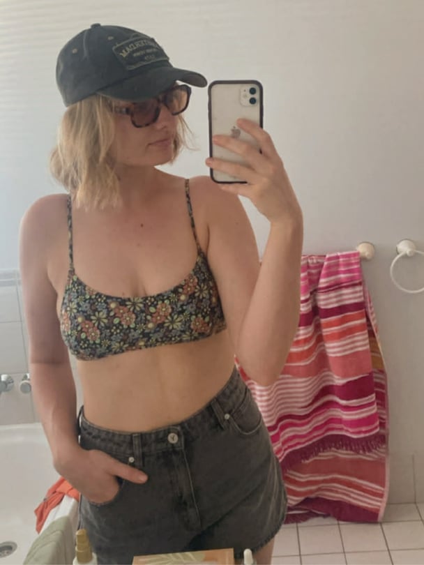 Natalie taking a mirror selfie in a bathroom, wearing a floral crop top, high-waisted shorts, sunglasses and a cap.