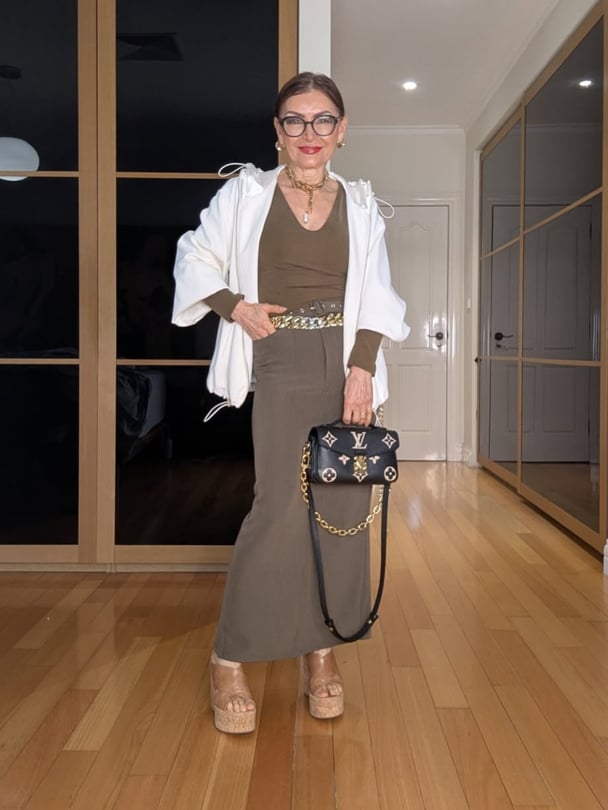 Faye in a brown maxi skirt with green long-sleeve top in a white jacket. She wears brown wedges and holds a Louis Vuitton bag.